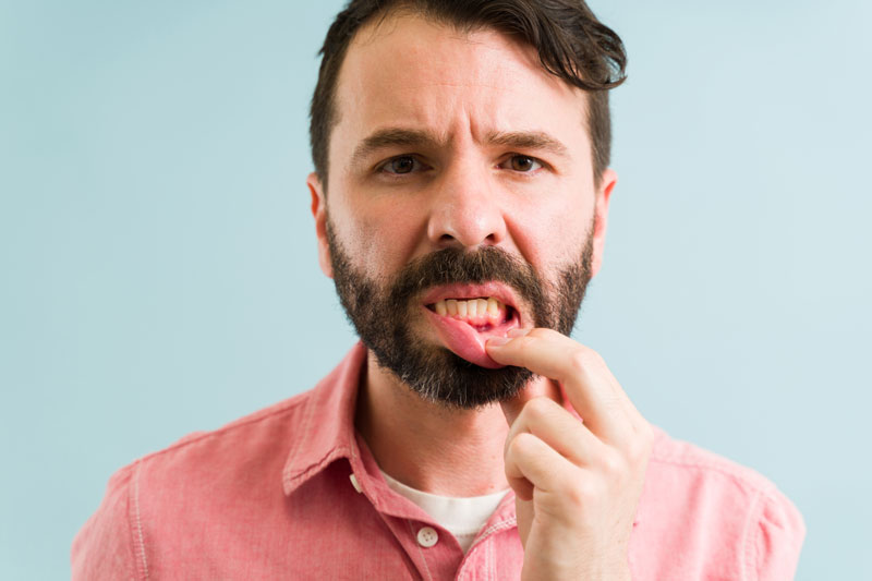 A man in a pink shirt pulls down his lower lip to expose his gums, looking concerned, likely inspecting for a dental or gum issue. Background is a soft blue.